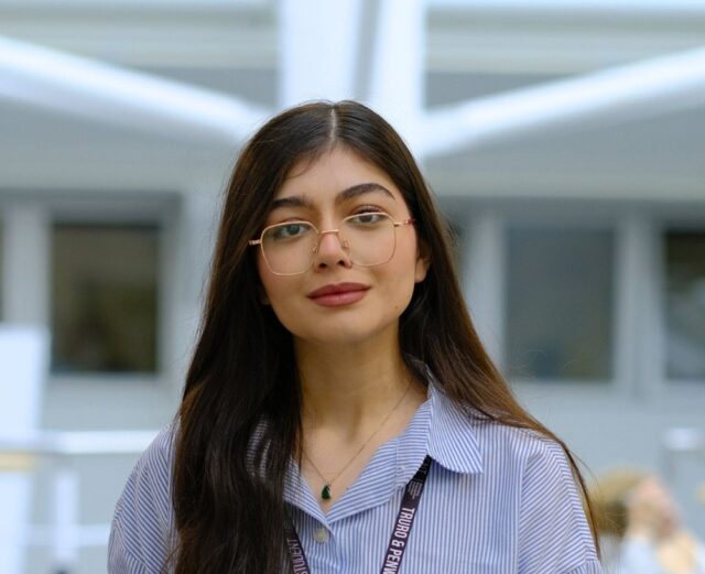 A student, Paria, stands looking at the camera. She wears a blue and white stripe shirt and has gold glasses.