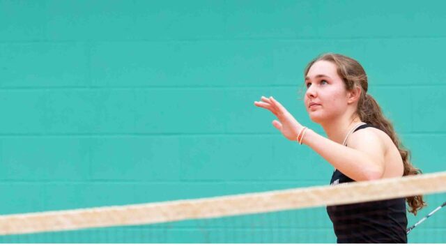 A student is playing badminton. She is wearing a black sports outfit and is stood against a blue wall. She looks up and readies herself to hit the shuttlecock.