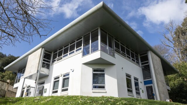 A photo of the Ginkgo building at Tregye. There is grass in the foreground, bare trees to the sides and a large white building in the centre.
