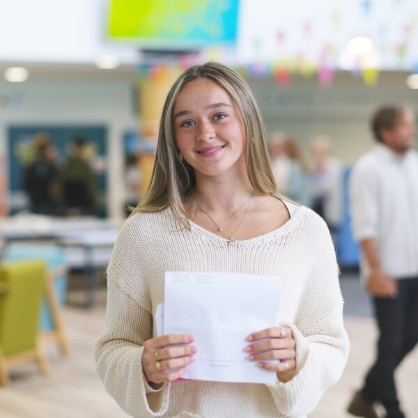A photo of Charlotte, she is wearing a cream jumper and is holding her results in front of her.