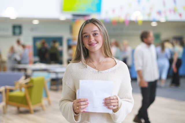 A photo of Charlotte, she is wearing a cream jumper and is holding her results in front of her.