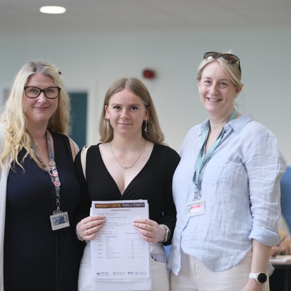 A photo of Hannah with two members of staff. She is holding her results up in front of her.