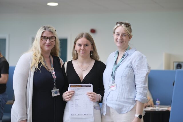 A photo of Hannah with two members of staff. She is holding her results up in front of her.