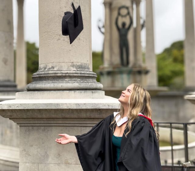 A photo of Emily, she is wearing a graduation gown and she is throwing her cap up in the air.