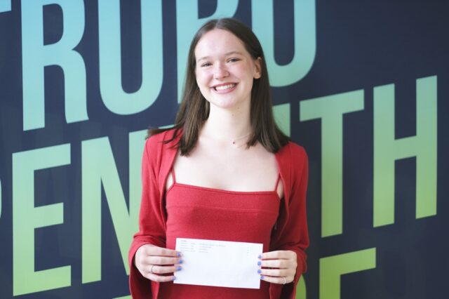 A photo of Mimi, she is wearing a red top and red cardigan and is standing in front of a Truro and Penwith College backdrop.