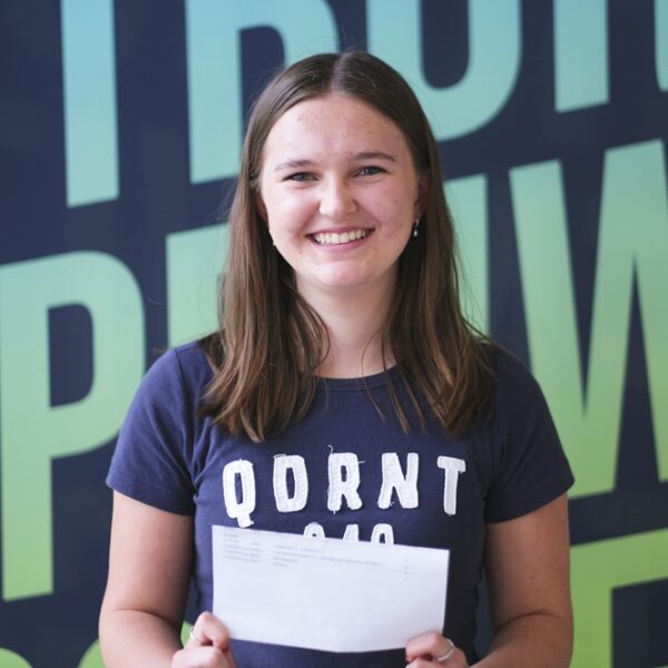 A photo of Poppy, she is wearing a navy t shirt and holding up her results in front of her. She has medium length brown hair and is smiling at the camera.