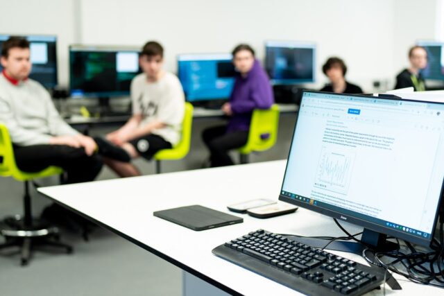 A close up of a computer on a table. Students can be seen in the background of the image.