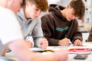 Two students can be seen working at desks with pens and paper.