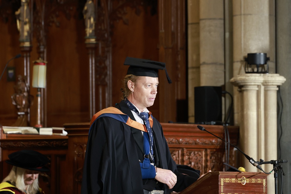 A photo of James McBlane stood at a lectern in graduation robes and his arm in a sling.