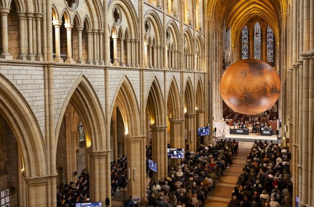 An aerial shot of Truro Cathedral during the University Centre graduation ceremony. People sat in seats and a large replica of Mars hangs over the stage.
