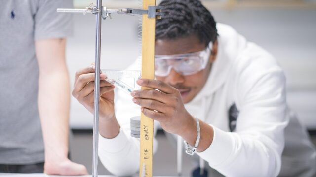 A student in a white jumper and wearing goggles holds a protrator against a long piece of wood.
