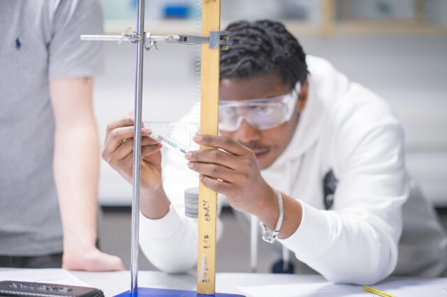 A student in a white jumper and wearing goggles holds a protrator against a long piece of wood.