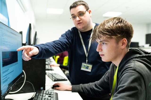 An image of a student sat at a computer with a member of staff. The tutor is pointing towards the computer screen.