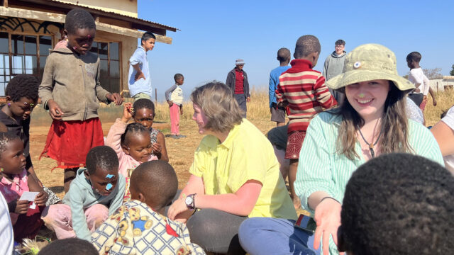 Students sit outside on the grass with children from Eswatini making crafts and playing with stickers.