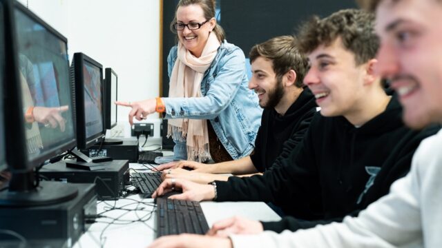 A group of HE students sit in front of computers as a tutor stands beside them and points towards one of the computer screens.