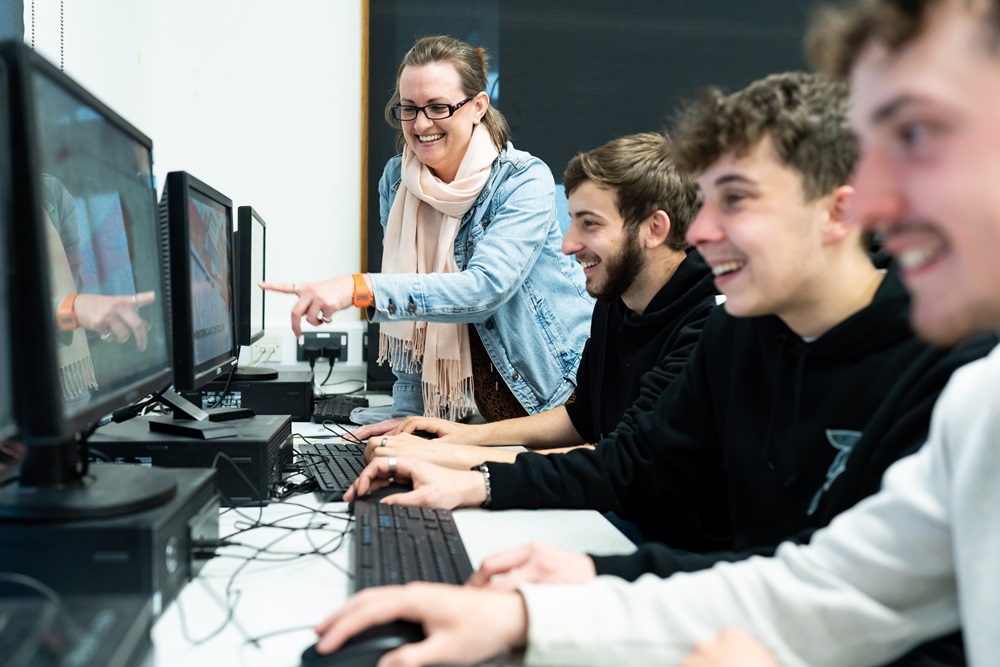 A group of HE students sit in front of computers as a tutor stands beside them and points towards one of the computer screens.