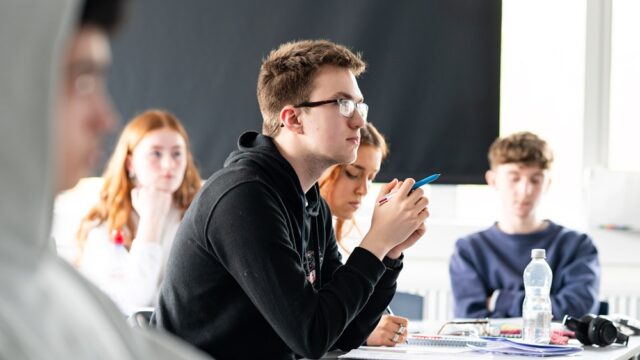 A group of students sit at classroom tables looking towards the front of the class. There are notepads and water bottles on the table.
