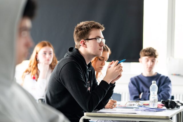 A group of students sit at classroom tables looking towards the front of the class. There are notepads and water bottles on the table.