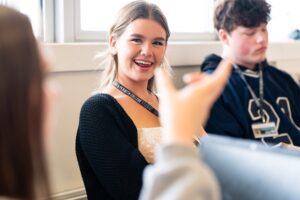 A photo of a student who is smiling at an unknown person. The unknown person is gesturing with their hand.