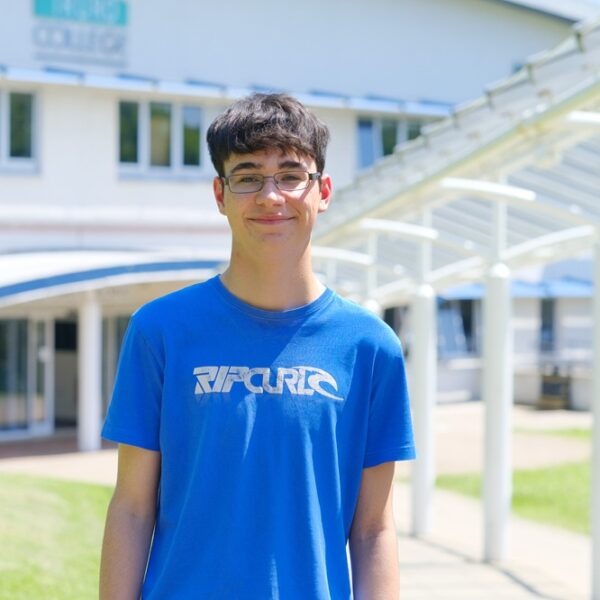 A photo of Nicholas stood outside the Helford building. He is wearing a blue t-shirt and smiling at the camera.