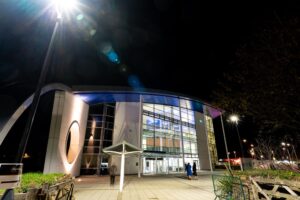 An exterior image of the Fal building at night time. The building is lit up from within and there is a streetlight in the top left corner that is producing a light flash.