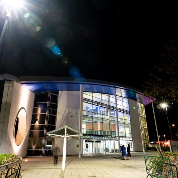 An exterior image of the Fal building at night time. The building is lit up from within and there is a streetlight in the top left corner that is producing a light flash.
