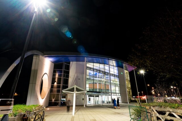 An exterior image of the Fal building at night time. The building is lit up from within and there is a streetlight in the top left corner that is producing a light flash.