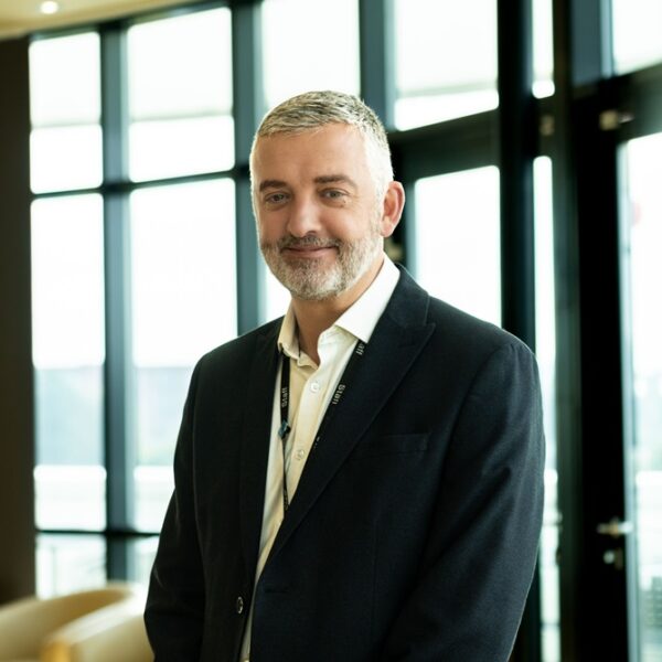 A photo of Carl Riding. He is wearing a suit and a lanyard and smiles at the camera.