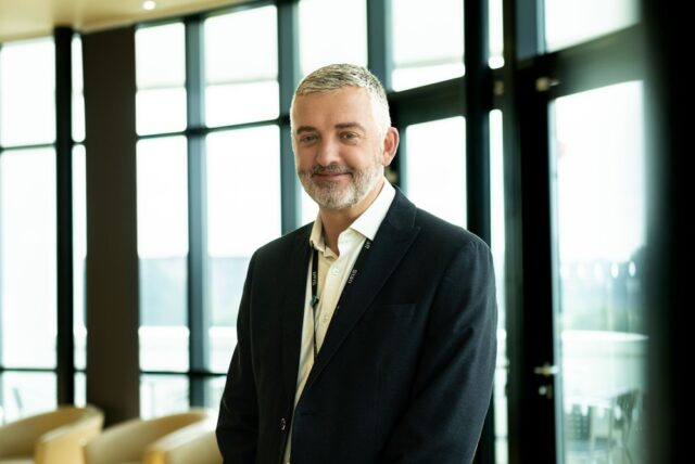 A photo of Carl Riding. He is wearing a suit and a lanyard and smiles at the camera.