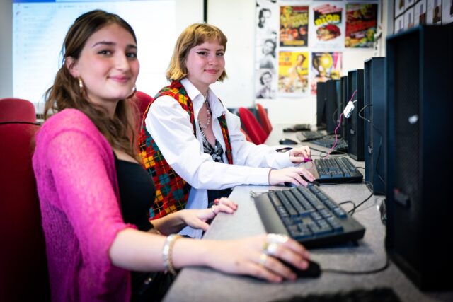 An image of two girls sat in a media class working on computers. They turn and smile at the camera.
