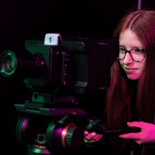 A girl looks through the lens of a large film camera. She has long red hair and wears glasses.