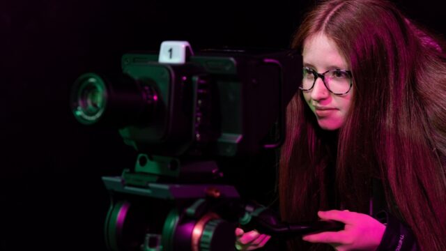 A girl looks through the lens of a large film camera. She has long red hair and wears glasses.
