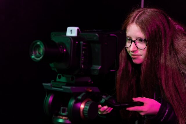 A girl looks through the lens of a large film camera. She has long red hair and wears glasses.