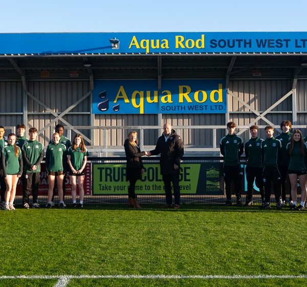 A football team stand on a football pitch. Two people in coats stand shaking hands.