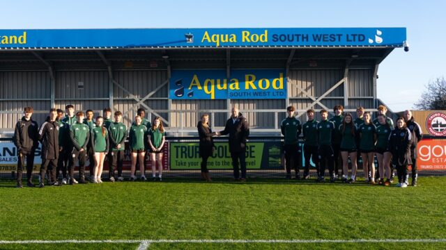 A football team stand on a football pitch. Two people in coats stand shaking hands.