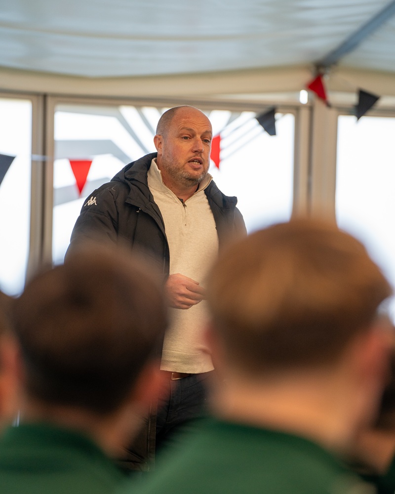 An image of a man stood wearing a fleece and jacket speaking to a crowd of people