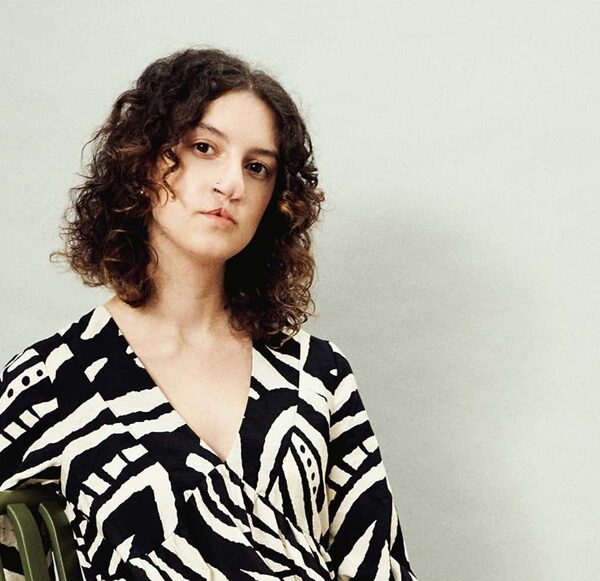 A girl with curly dark hair and a black and white dress on sits in a chair and tilts her head at the camera.
