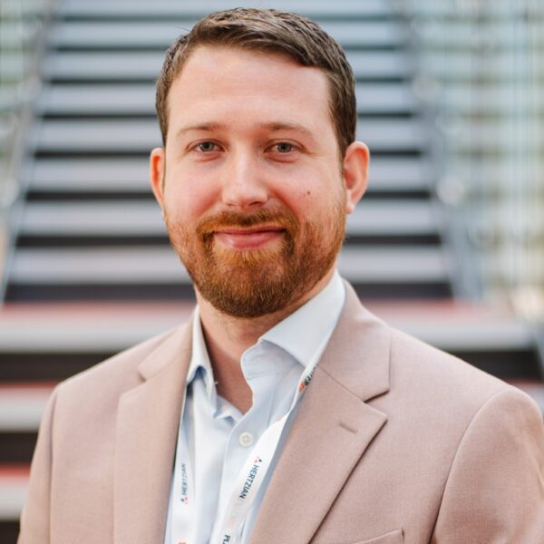 A photo of Christopher Weavill. He is wearing a light coloured suit, a white shirt and a lanyard that says Hertzian on it.