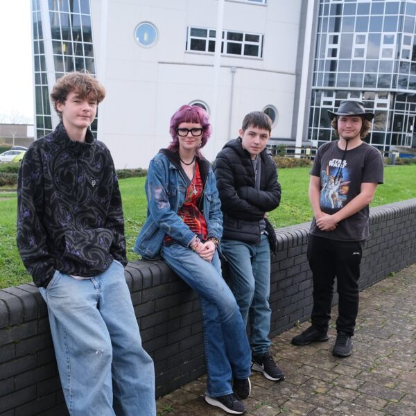 Four students lean against a low wall outside Penwith College.