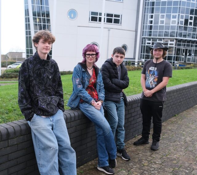 Four students lean against a low wall outside Penwith College.