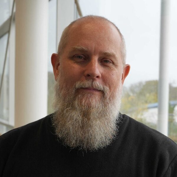 A head and shoulders photo of a man who has a beard and is wearing a dark top.