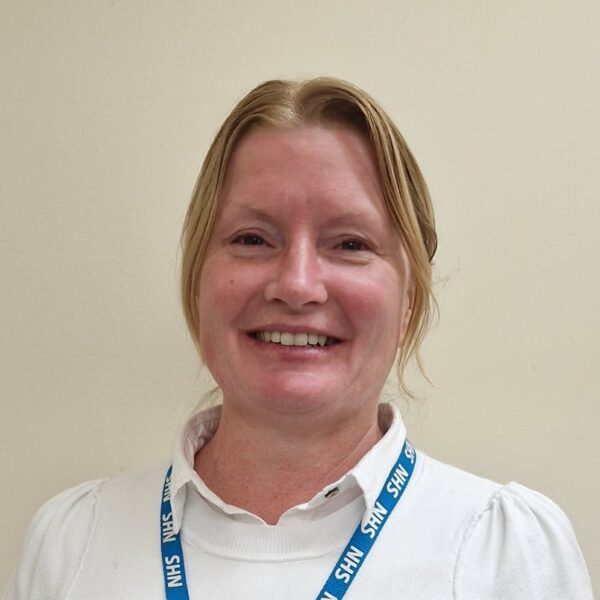 A head and shoulders photo of a woman with blonde hair who is wearing a white top and blue NHS lanyard.