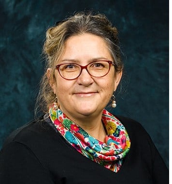 A portrait of Sarah Sanderson. She is wearing a black top with a colourful scarf and she is sat against a dark background.