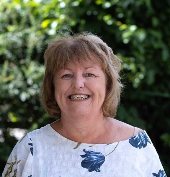 A photo of Tamsin Merrit, she is stood outside and is wearing a white top with blue flowers on it.