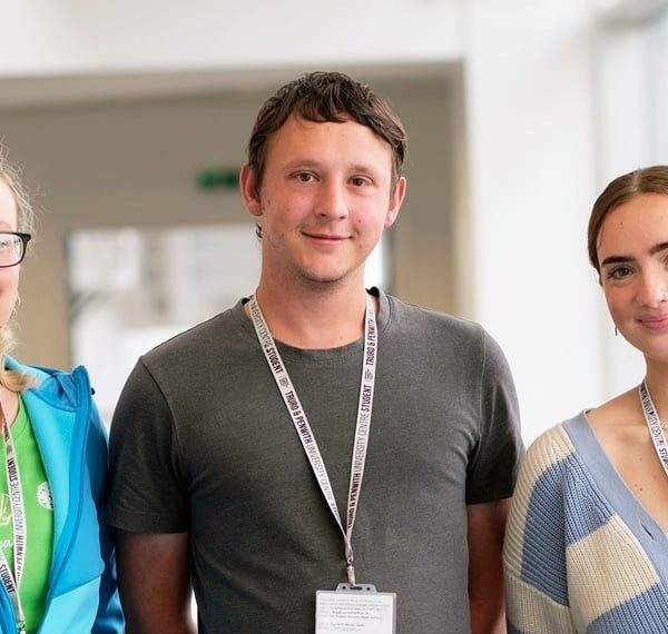 Three students smile at the camera. They are wearing colourful clothing and each of them wears a white lanyard with a student card attached.