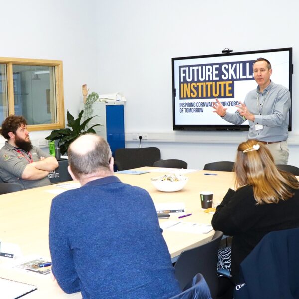 A tutor stands in front of a small group of students. They are all sat at a conference table.