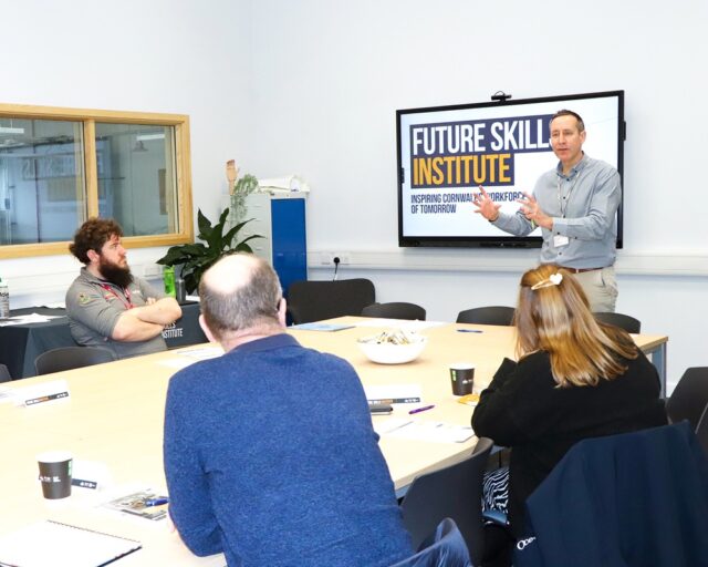 A tutor stands in front of a small group of students. They are all sat at a conference table.