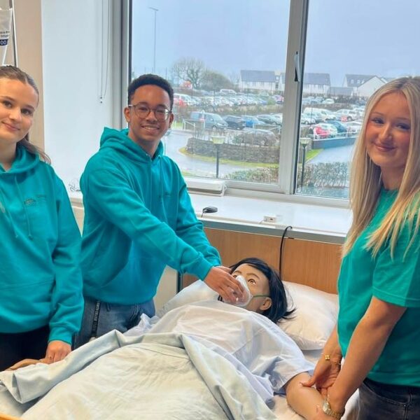 Three students stand around a dummy in a hospital bed. They are taking the dummy's pulse and holding an oxygen mask to its face.