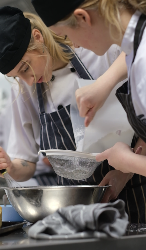 Two students in chefs whites use a sieve over a metal bowl.