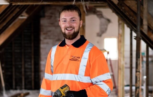A construction apprentice holding an electric drill in the loft of a newly constructed building.